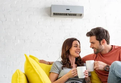 A man and a woman sitting on a couch with cups of coffee with a wall-mounted AC unit behind them.