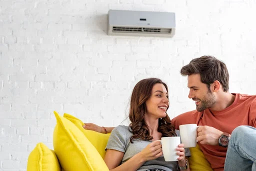 A man and a woman sitting on a couch with cups of coffee with a wall-mounted AC unit behind them.