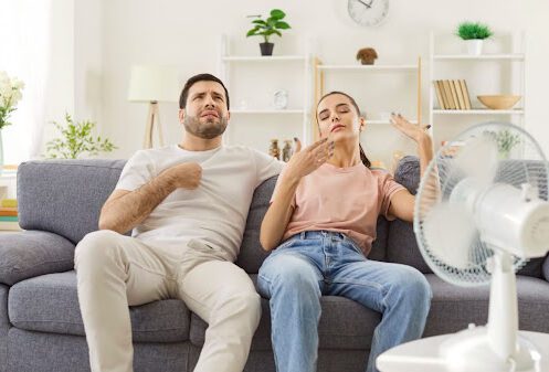 A man and a woman sitting in front of a fan and fanning themselves in a warm home.