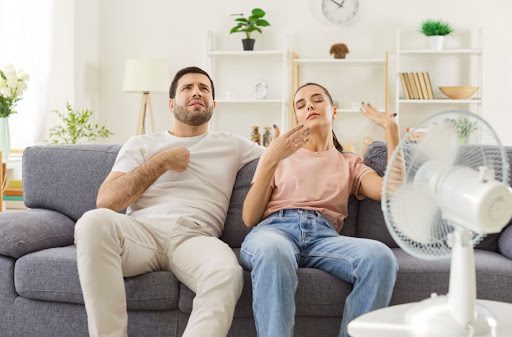 A man and a woman sitting in front of a fan and fanning themselves in a warm home.