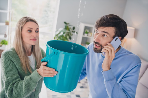 A woman holding a bucket to catch a leak from a home's ceiling while a man calls for help.