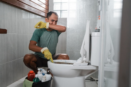 A man covering his nose due to a smelly toilet; he has cleaning supplies with him.
